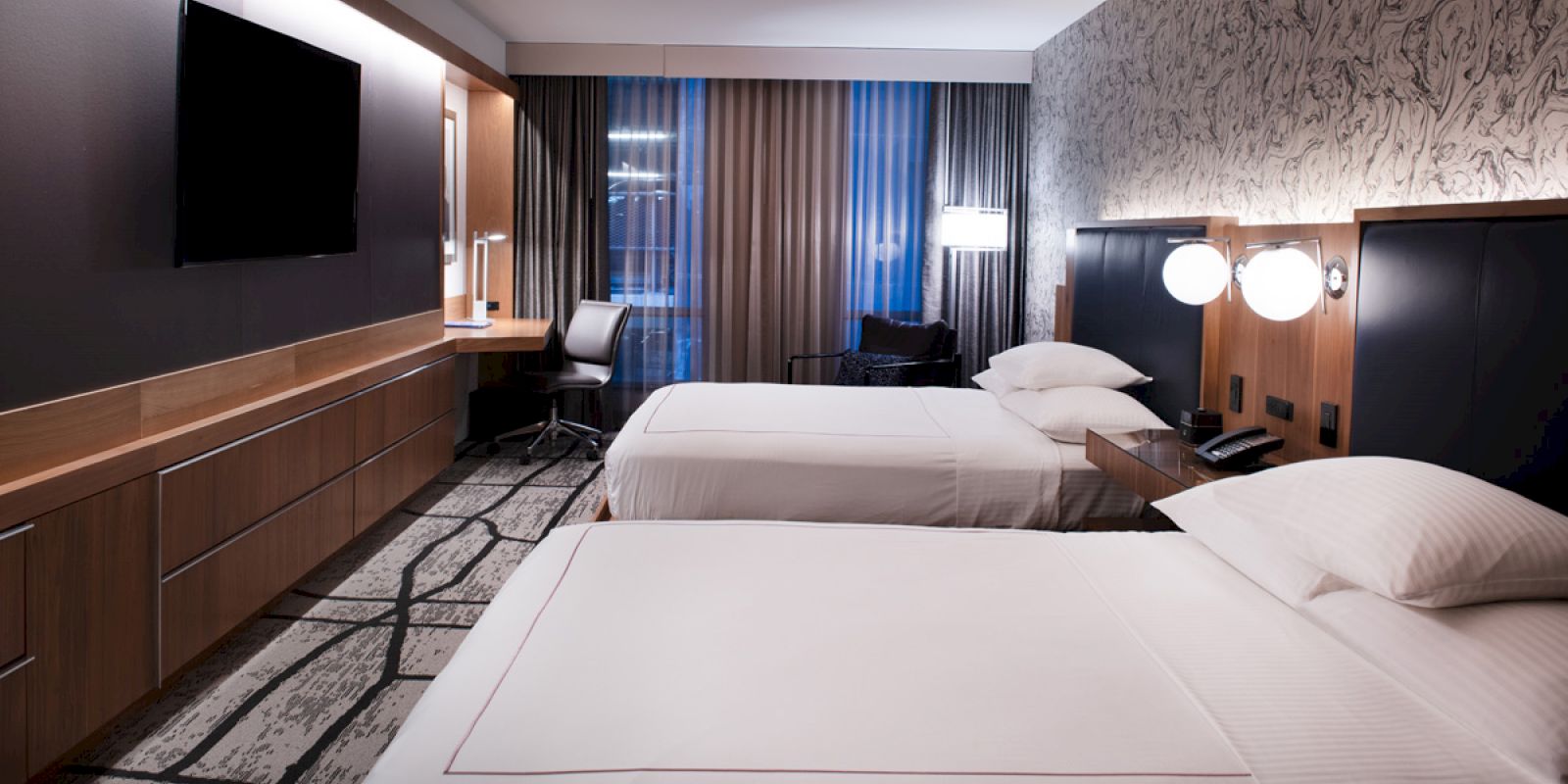 A modern hotel room with two beds, a flat-screen TV, a desk, chairs, and stylish decor featuring wallpaper and curtains, illuminated by lamps.