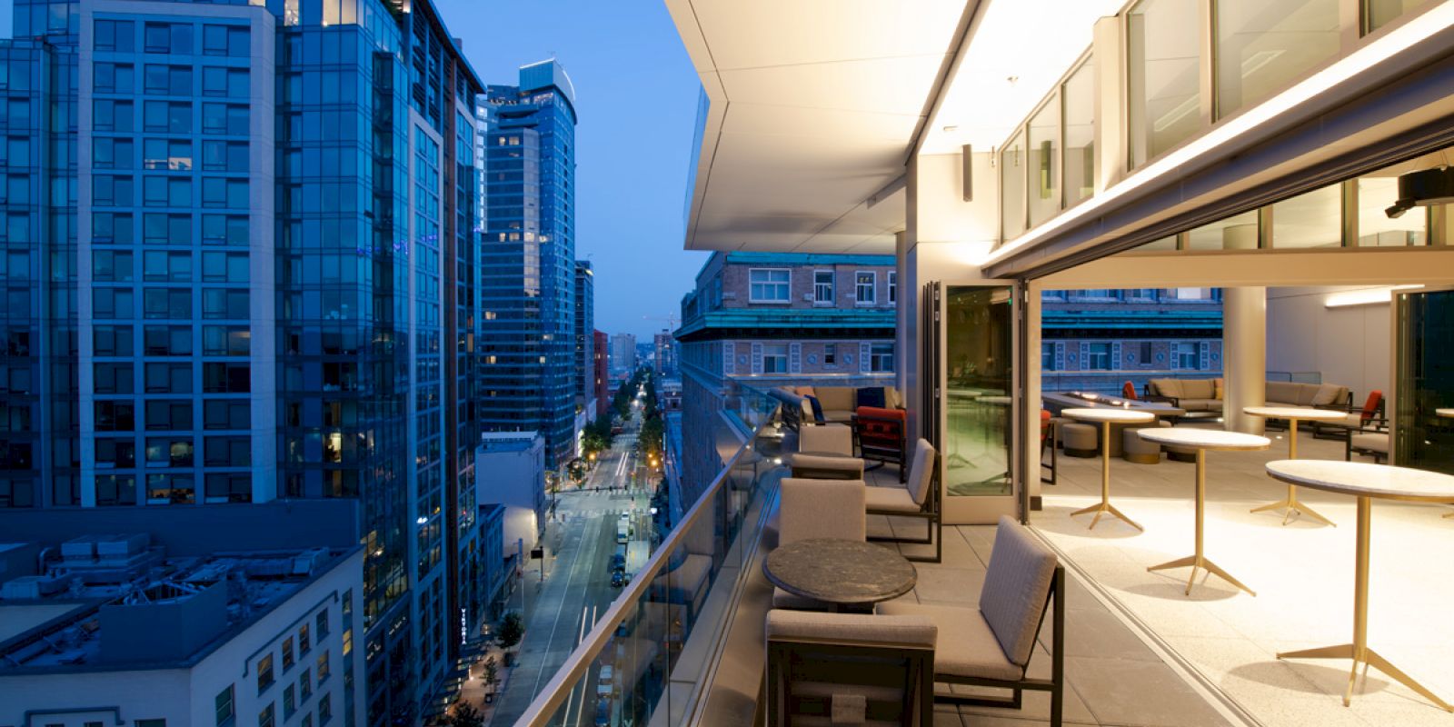 A modern cityscape with tall buildings at dusk, featuring a spacious balcony with outdoor seating and tables, overlooking a busy street below.