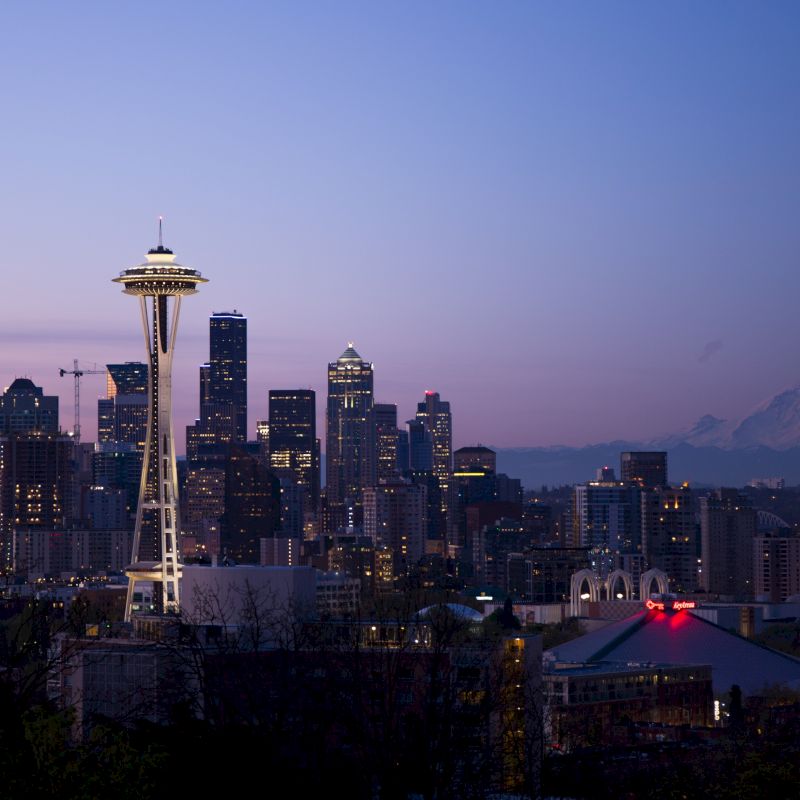 A city skyline at dusk featuring the Space Needle with illuminated buildings and Mount Rainier in the distance under a purple sky, ending the sentence.