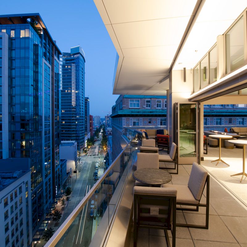 A modern balcony at dusk, overlooking a city street with tall buildings, furnished with tables, chairs, and lit by warm indoor lighting.