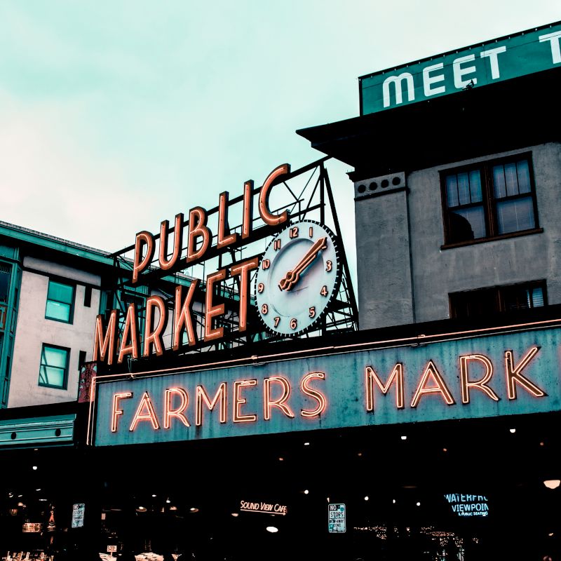 A neon sign reads "Public Market" and "Farmers Market" with a clock in the middle, set against buildings with various signage.