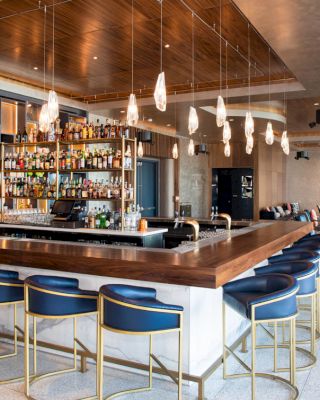 A modern bar with a wooden counter, blue bar stools, hanging lights, and a well-stocked liquor display, located in a chic lounge area.