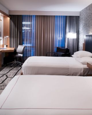 The image shows a modern hotel room with two neatly made beds, a desk, a chair, a TV, and contemporary lighting fixtures.