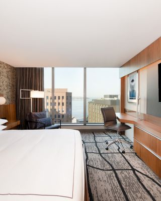A modern hotel room with a large bed, flat-screen TV, desk, and a window offering a city view. The decor features wood paneling and contemporary design.
