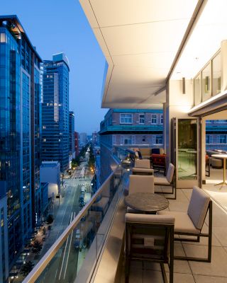 The image features a modern cityscape with tall buildings at dusk and a spacious, furnished balcony overlooking the street below.