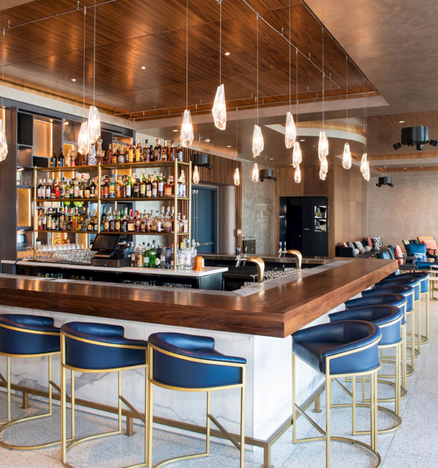 A modern bar with elegant seating, contemporary lighting fixtures, a well-stocked shelf of bottles, and a cozy seating area in the background.