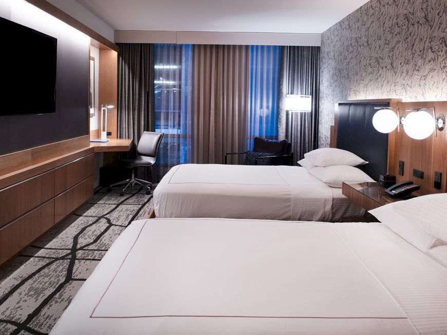 A modern hotel room with two neatly made beds, a wall-mounted TV, a desk with a chair, and a stylish decor featuring patterned wallpaper and curtains.