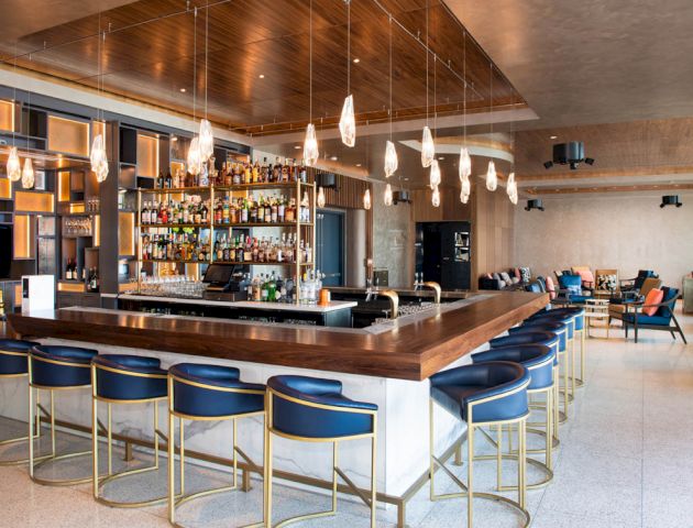 The image shows a modern bar with a wooden counter, high chairs, hanging lights, and stocked shelves, set in a spacious, elegant environment.