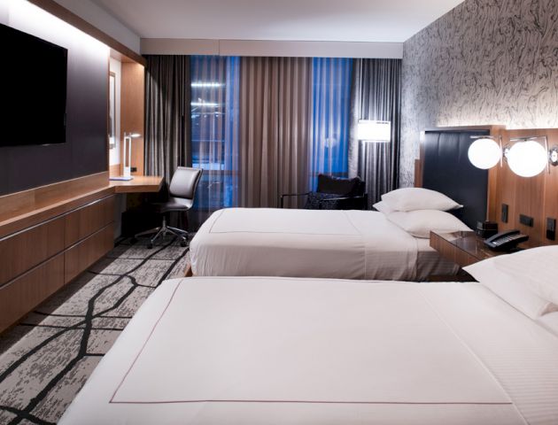 A modern hotel room with two double beds, a flat-screen TV, a work desk, and contemporary decor with patterned wallpaper and curtains.