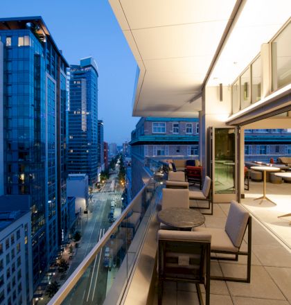 The image shows a modern apartment balcony with a seating area overlooking a city street lined with high-rise buildings at dusk.