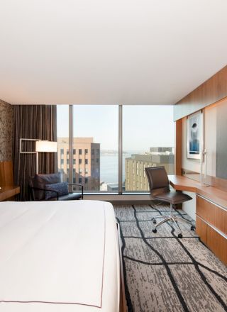 A modern hotel room with a large bed, desk, chair, wall-mounted TV, and window overlooking city buildings and water, with contemporary decor.