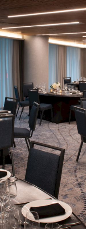 The image shows a modern banquet hall setup with round tables, chairs, and elegant table settings including glasses, cutlery, and napkins on each table.