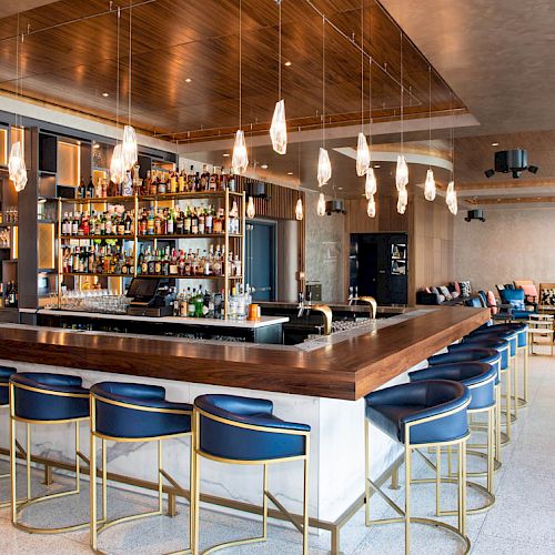 The image shows a modern bar with a large selection of bottles, stylish lighting, and a seating area with blue stools and lounge chairs in the background.
