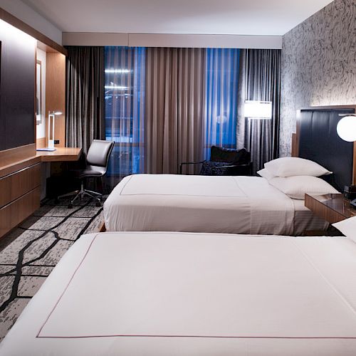 A modern hotel room with twin beds, a wall-mounted TV, a desk, and a chair. The room features contemporary decor and ample lighting.