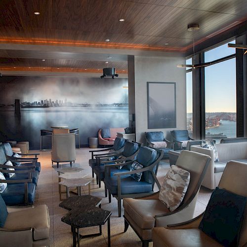 A modern lounge with leather chairs, a wall mural of a city skyline, large windows with a coastal view, and a Ferris wheel visible outside.