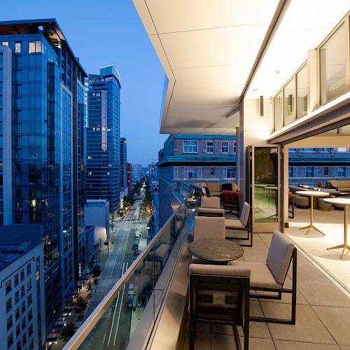 A modern rooftop patio at dusk, featuring seating areas and tables, with a view of city high-rise buildings and illuminated streets below.