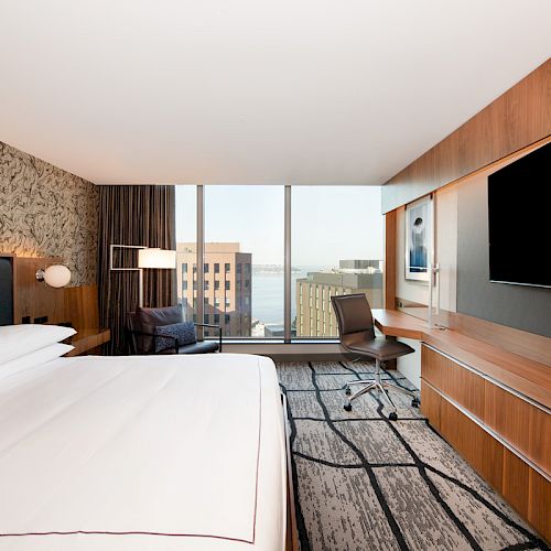 A modern hotel room with a large bed, wall-mounted TV, work desk, armchair, and a window offering a city view.