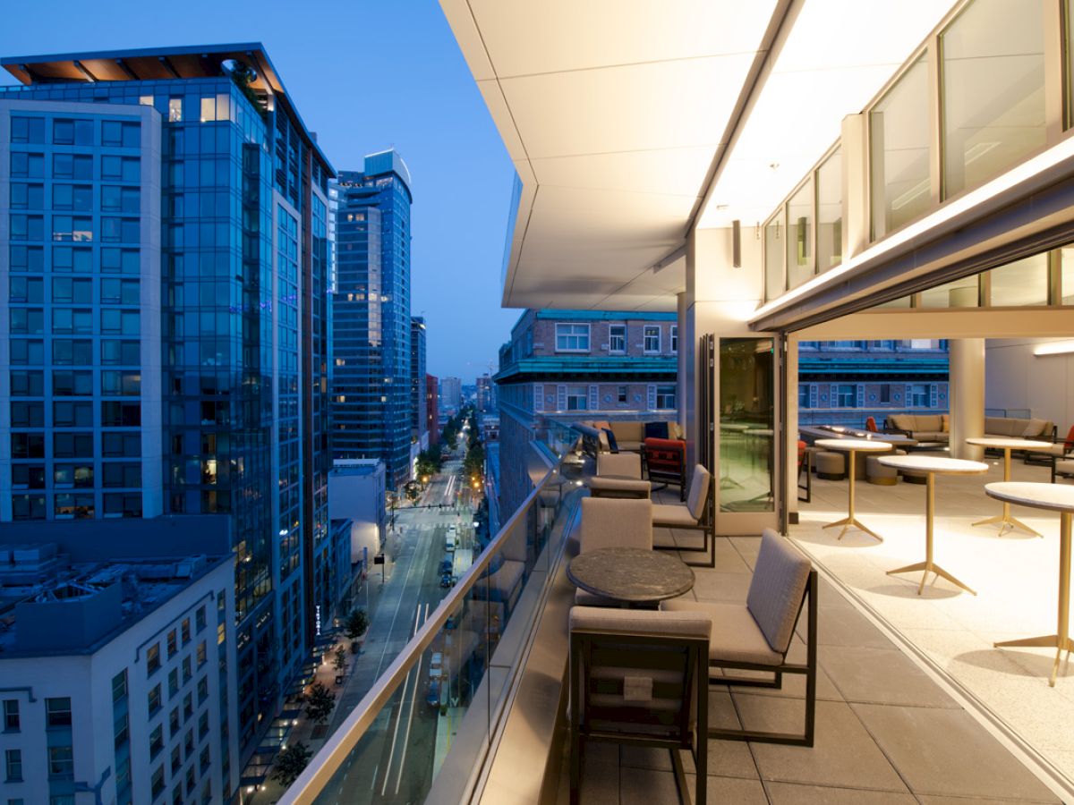 A modern balcony overlooks a cityscape during the evening, with tables and chairs arranged for outdoor seating, and buildings visible in the background.