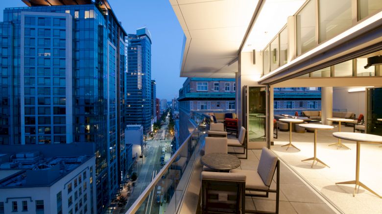 A modern balcony overlooks a cityscape during the evening, with tables and chairs arranged for outdoor seating, and buildings visible in the background.