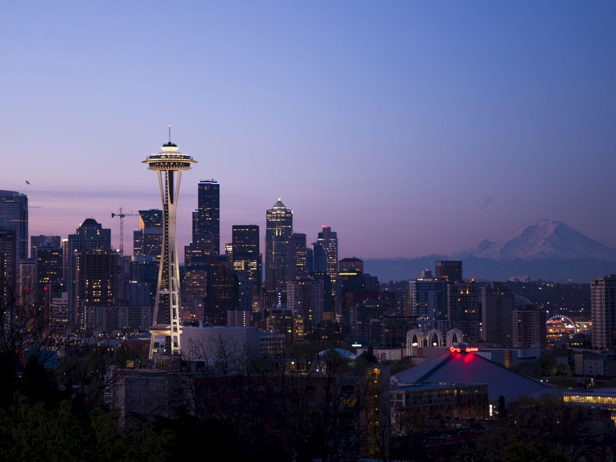 The image shows the Seattle skyline at dusk, prominently featuring the Space Needle with Mount Rainier in the background and city lights beginning to illuminate.