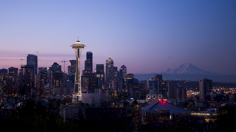 The image shows the Seattle skyline at dusk, prominently featuring the Space Needle with Mount Rainier in the background and city lights beginning to illuminate.