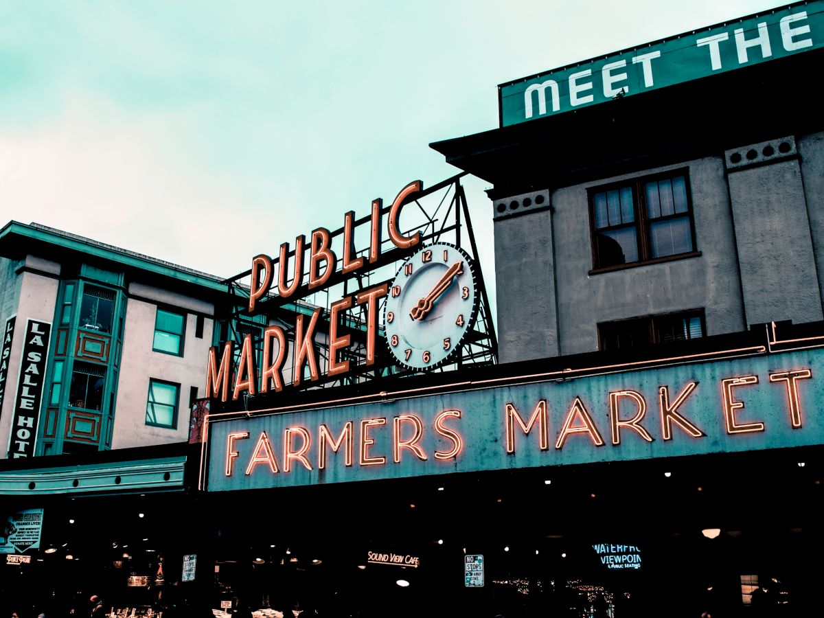 The image shows a neon sign for a "Public Market" and "Farmers Market," with a clock in the middle, situated in an urban setting.