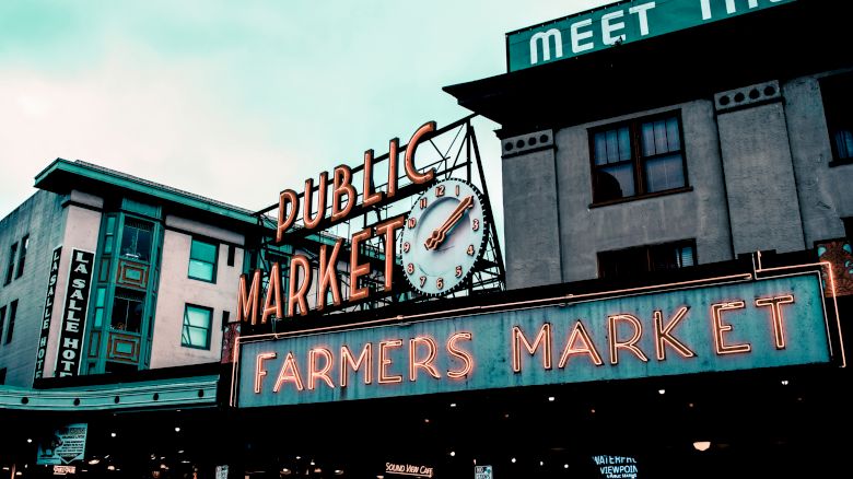 The image shows a neon sign for a "Public Market" and "Farmers Market," with a clock in the middle, situated in an urban setting.