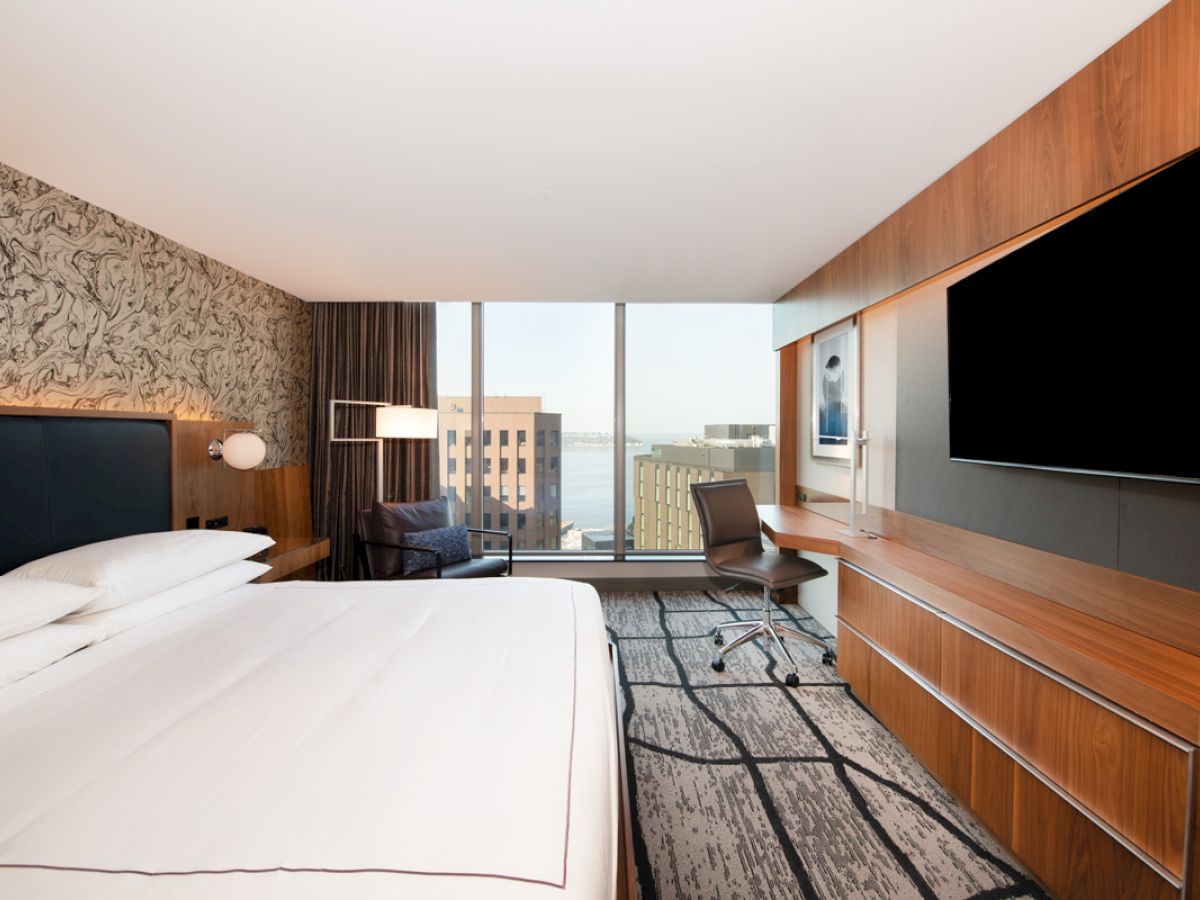A modern hotel room with a large bed, wall-mounted TV, desk, and a view of city buildings through a floor-to-ceiling window ends the sentence.