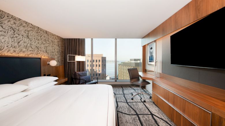 A modern hotel room with a large bed, wall-mounted TV, desk, and a view of city buildings through a floor-to-ceiling window ends the sentence.