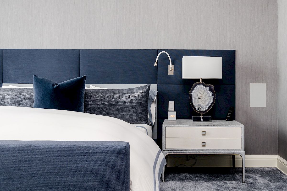 A neatly arranged bedroom with a blue headboard, white bed, blue cushions, a bedside table with a lamp, and a white drawer for storage.