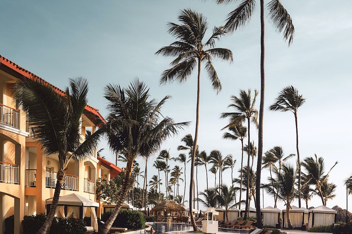 A tropical resort with a swimming pool, sun loungers, palm trees, and a building with balconies against a clear sky are shown.