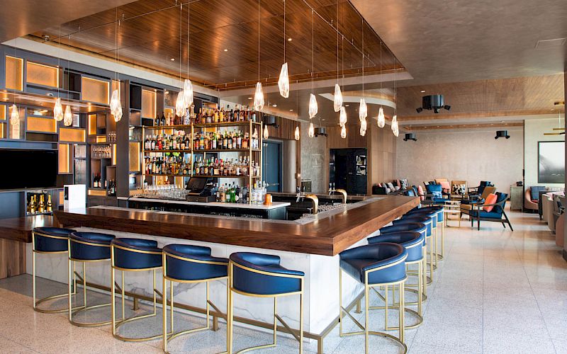The image shows a modern bar with a wooden counter, blue stools, a fully stocked back bar, and contemporary lighting. There is seating in the background.