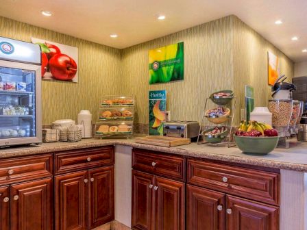 A breakfast buffet area with a variety of foods and beverages, including fruits, pastries, cereal, a toaster, coffee maker, and refrigerator.